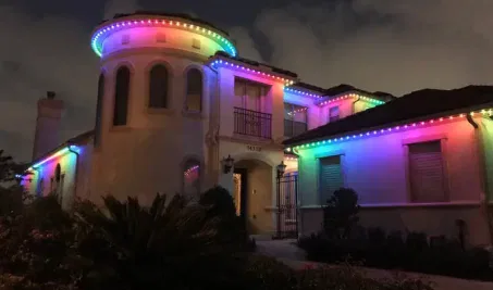 A large white house at night is decorated with bright, multi-colored LED lights outlining the roof, creating a festive and colorful display against a cloudy sky.