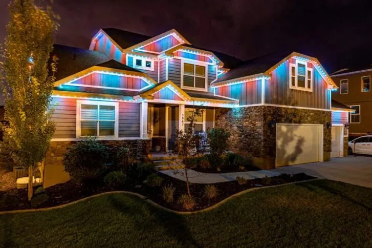 A two-story house at night is decorated with colorful, glowing lights along the roofline, illuminating the exterior and yard. The house has a stone and siding facade, multiple windows, and a neatly landscaped lawn.