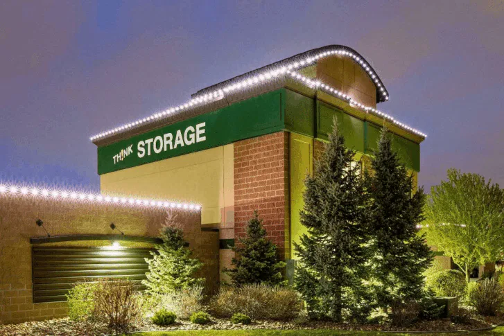 A self-storage building with Think Storage on a green sign, outlined with white lights at the roofline. Evergreen trees and shrubs are landscaped in front, and the sky has a soft purple hue.