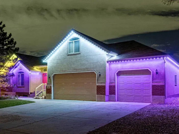A house at night with white and purple LED lights outlining the roof and garage, illuminating the driveway and yard, with trees and a cloudy sky in the background.